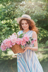 Portrait of young redhead curly woman in straw hat and linen stripe dress with a basket and a pink  peonies bouquet in the garden
