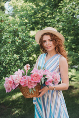 Fototapeta premium Portrait of young redhead curly woman in straw hat and linen stripe dress with a basket and a pink peonies bouquet in the garden