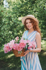 Portrait of young redhead curly woman in straw hat and linen stripe dress with a basket and a pink  peonies bouquet in the garden