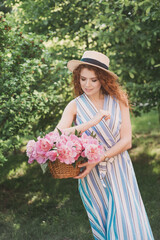 Portrait of young redhead curly woman in straw hat and linen stripe dress with a basket and a pink  peonies bouquet in the garden