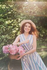 Portrait of young redhead curly woman in straw hat and linen stripe dress with a basket and a pink  peonies bouquet in the garden