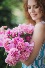Portrait of young redhead curly woman in straw hat and linen stripe dress with a basket and a pink peonies bouquet in the garden. No face closeup