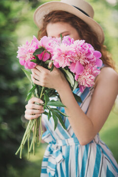 Portrait of young redhead curly woman in straw hat and linen stripe dress with a basket and a pink  peonies bouquet in the garden
