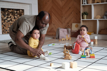 Mature African American man spending time at home with his twin daughters watching them playing with toys and taking care of them