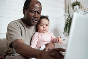 Obraz premium Portrait of modern mature African American man sitting on sofa with baby daughter on his lap working on laptop