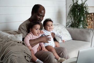 Fototapeta premium Portrait of mature African American father sitting on sofa in living room with twin baby daughters on his lap watching cartoons in Internet