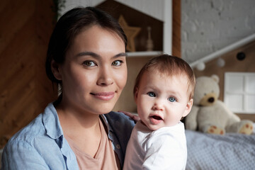 Close-up portrait of beautiful young mother spending time at home holding baby daughter in arms looking at camera