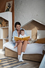 Modern happy young mother sitting in cozy kids room with baby daughter on lap reading fairy-tale