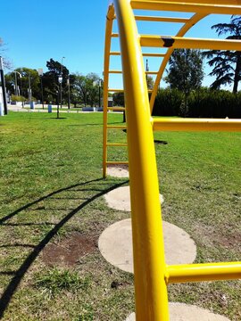 Par&aacute;bola de tubo de acero amarillo con escalones para juegos infantiles y ejercicios en el patio de recreo, forma una sombra curva en el c&eacute;sped con un fondo contrastante de &aacute;rboles y cielo azul