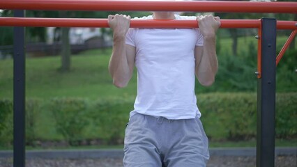 Motivated focused man in wheelchair doing pull ups in slow motion outdoors. Front view portrait of inspired Caucasian concentrated sportsman with disability training on fitness equipment in park