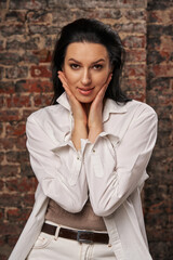 a beautiful woman with dark hair and in a white shirt holds her face with her palms and looks in surprise at the frame against the background of a brick wall