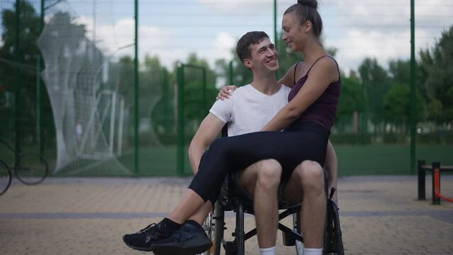 Front view happy Caucasian man in wheelchair rolling in slow motion as smiling woman sitting on lap kissing partner. Loving boyfriend and girlfriend dating outdoors enjoying leisure. Love lifestyle