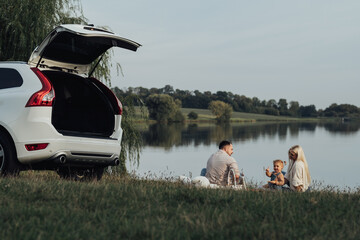 Happy Young Family Enjoying Picnic Time Outside the City, Mother and Father with Their Daughter Sitting Outdoors Near Their SUV Car, Road Trip Concept