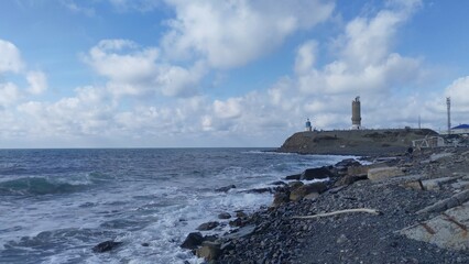 Foamy sea waves roll on the rocky shore. On a hilly promontory stand a lighthouse, a chapel and a metal communication tower. The sky above the sea is blue with clouds