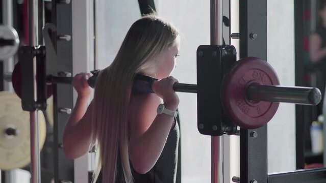 Back view of blonde woman does squats with barbell on smith machine at gym