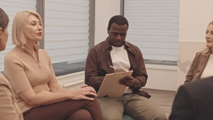Medium slowmo of group of mature men and women and African American male psychologist sitting in circle while discussing mental problems during group therapy session - Powered by Adobe