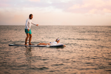 Happy father and daughter surfing on a sup board and have a funny time. Sunset ocean at the background. Family summer vacations