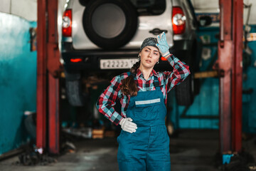 A young female mechanic in uniform wipes the sweat from her forehead wearily. In the background-a car on a lift. Stress in the workplace
