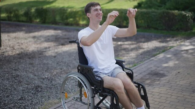 Portrait of young cheerful Caucasian man in wheelchair catching passing basketball ball in slow motion outdoors. Smiling positive person with disability enjoying sport exercising on sunny day