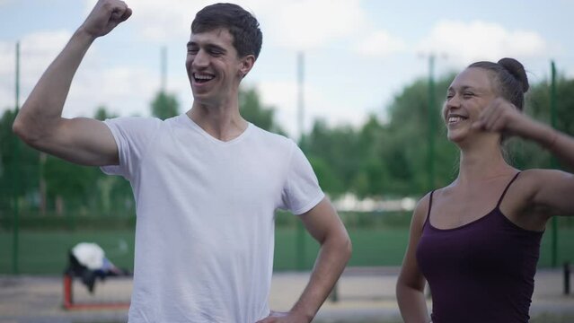 Positive Caucasian young man and woman gesturing high-five showing strength gesture smiling looking at camera. Confident sportive friends posing in slow motion outdoors on sports ground in park