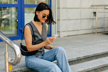 Stylish woman in grey vest and jeans with bag is messaging on phone. 