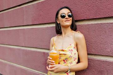 Glad girl in summer stylish outfit having fun outdoor waiting friend. Portrait of pretty female model in yellow dress is laughing while drinking lemonade on blur street background.