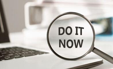 DO IT NOW - text on magnifying glass lying on laptop on office table.