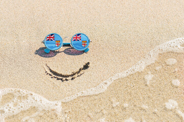 A painted smile on the sand and sunglasses with the flag of Fiji. The concept of a positive and successful holiday in the resort of Fiji.