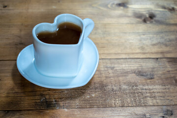A white cup of black strong coffee in a heart-shaped mug and plate in the shape of a heart. Hot drink on brown wood background.