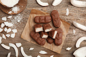 Delicious milk chocolate candy bars with coconut filling on wooden table, flat lay