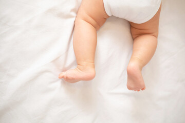 The legs of the baby in a white bodysuit on the white background of the bed. Light colors. Lifestyle.