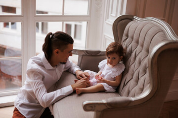 Handsome happy father playing with legs of little daughter on beige sofa indoors. Young man in white shirt with his 1-year-old daughter in white dress, holding her legs. family tenderness