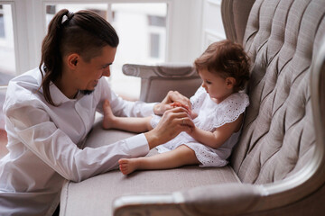 Handsome happy father playing with legs of little daughter on beige sofa indoors. Young man in white shirt with his 1-year-old daughter in white dress, holding her legs. family tenderness