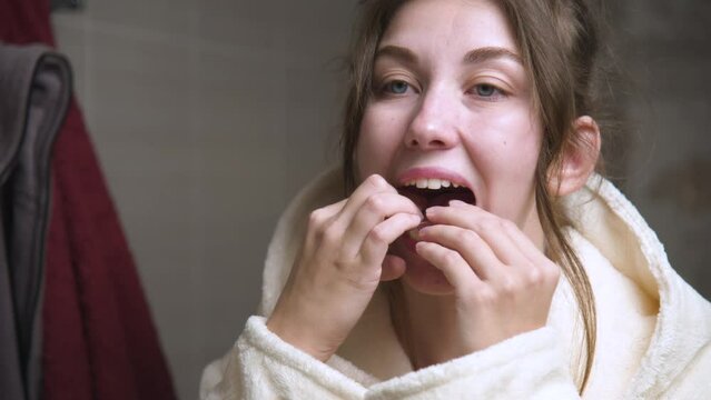 Attractive young Caucasian woman in a bathrobe standing in the bathroom installs a polymer aligner on her lower jaw. Oral care and bite restoration and teeth alignment