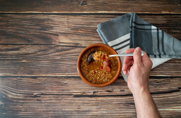 top view of a hand holding a spoon with lentils from an earthenware plate on a wooden table