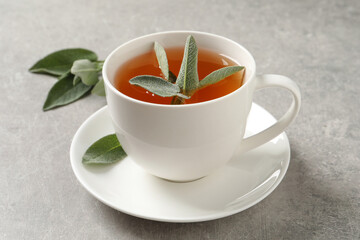 Cup of aromatic sage tea and fresh leaves on grey table