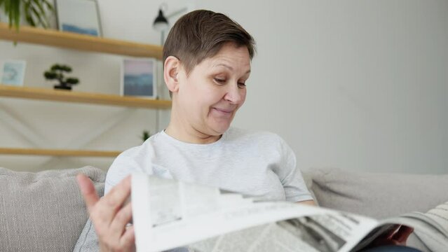A close-up view of a beautiful mature woman is reading a newspaper sitting on sofa at home in the morning. Portrait of senior woman reading newspaper