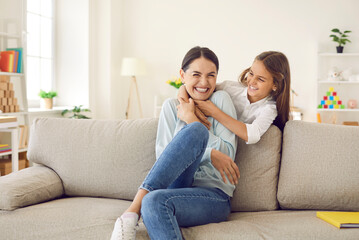 Happy mother hugging together and having fun with her teenage daughter at home on weekend. Cheerful child tickles his positive young mother sitting on sofa in living room. Family concept.