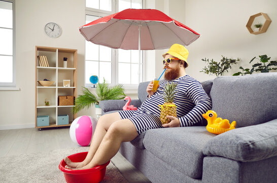 Funny man in living room at home imagines that he is resting on sea and sunbathing on beach. Chubby man wets his feet in plastic bowl and drinks cocktail while sitting on sofa under beach umbrella.