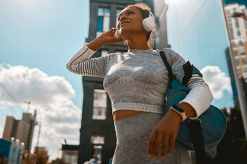 Happy sporty woman in headphones and sportswear smiling away standing on an urban street. Sports