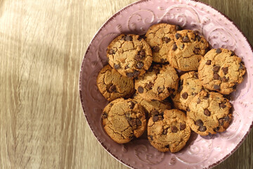 Pink plate filled with chocolate chip cookies on wooden table. Flat lay.