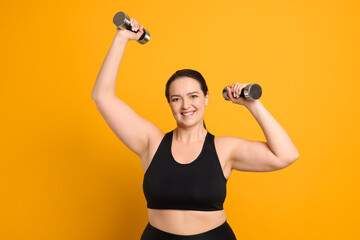 Happy overweight woman doing exercise with dumbbells on orange background