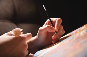 Close-up of a young female artist's hand with a thin brush painting a picture on canvas in a dark room. Shallow depth of field. high contrast