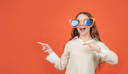 Happy girl child in big funny sunglasses pointing fingers aside brown background, copy space