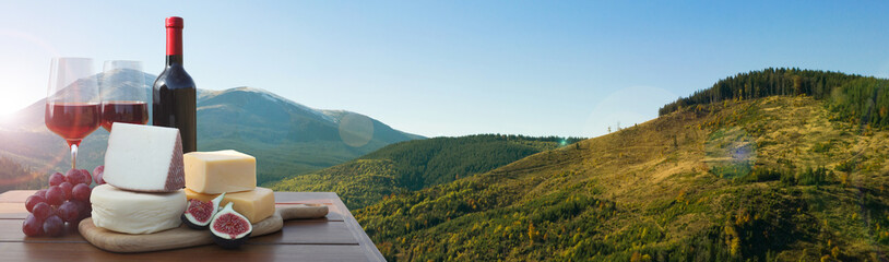 Tasty wine, grapes and cheeses on wooden table against beautiful mountain landscape, space for...