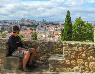 Lisbon, Portugal - July 07 2019: Castelo de Sao Jorge: caucasian man seated on the walls of the castle. He wears a hat, a black shirt and brown bermudas