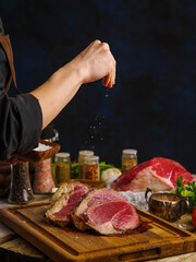 The chef prepares meat steaks on a dark background. Salt the cut pieces of meat. Lots of ingredients on the kitchen table. Restaurant, hotel, home cooking. Food background.