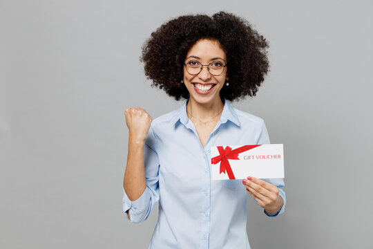 Young employee business corporate lawyer woman of African American ethnicity in shirt work in office hold gift certificate coupon voucher card for store do winner gesture isolated on grey background.