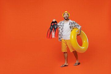 Full body young tourist fun man in beach shirt hat hold inflatable ring flippers walking go look camera isolated on plain orange background studio portrait. Summer vacation sea rest sun tan concept. © ViDi Studio