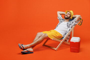 Young minded happy cool tourist man in beach shirt hat lie on deckchair near fridge hold hands behind neck isolated on plain orange background studio portrait Summer vacation sea rest sun tan concept - obrazy, fototapety, plakaty
